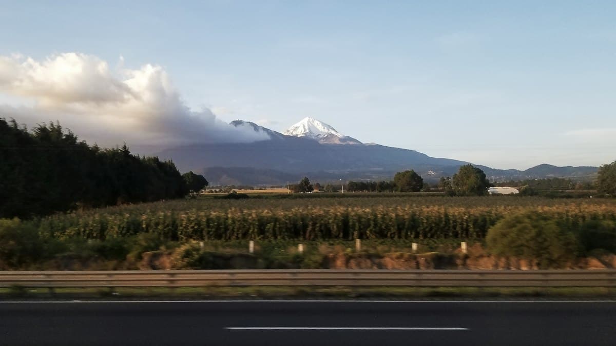 Autopista mexicana vista desde el coche, con un campo de maíz y un volcán nevado al fondo